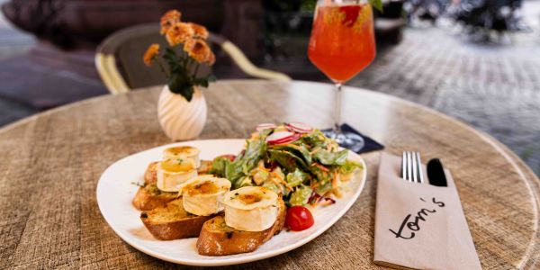 Ein runder Tisch im Außenbereich mit einem Teller gratiniertem Ziegenkäse auf Baguette mit Salat, einer kleinen Blumenvase und einem orangefarbenen Cocktail.