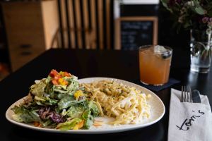 Ein Teller mit cremiger Pasta und einem großen, frischen Beilagensalat auf einem Restauranttisch. Im Hintergrund steht ein fruchtiger Cocktail.