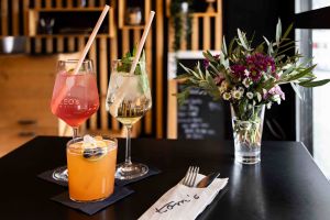 Drei sommerliche Cocktails auf einem dunklen Tisch, dekoriert mit Beeren, Minze und Strohhalmen. Daneben liegt Besteck in einer Serviette mit der Aufschrift "tom's".