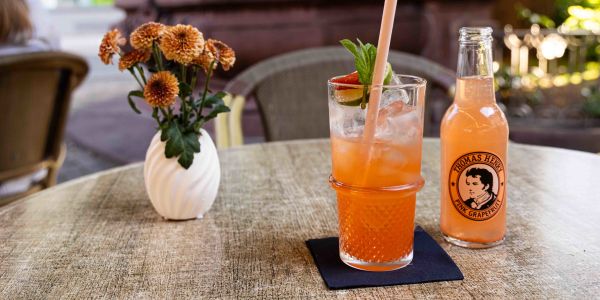 Ein runder Tisch im Außenbereich mit einer kleinen Blumenvase und einer Flasche Thomas Henry Pink Grapefruit neben einem Paloma-Highball mit Strohhalm, Limette und Minze auf einer dunklen Serviette.