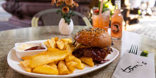 Ein runder Tisch im Außenbereich mit Burger und Pommes auf einem Teller samt Ketchup und Mayo, einer kleinen Blumenvase und einer Limonade im Hintergrund.