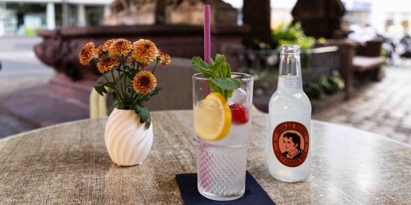 Ein runder Tisch im Außenbereich mit einer kleinen Blumenvase, einem Glas mit Zitrone, Himbeere und Minze auf einer dunklen Serviette sowie einer Flasche Thomas Henry Spicy Ginger Beer.