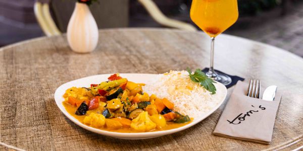 Ein runder Tisch im Außenbereich mit einem Teller Gemüse-Curry mit Reis, einer Vase mit roten Rosen und einem gelb-orangefarbenen Cocktail.