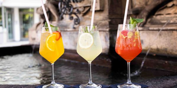Drei farbenfrohe Cocktails in Weingläsern mit Strohhalmen nebeneinander auf dem Rand eines Steinbrunnens.