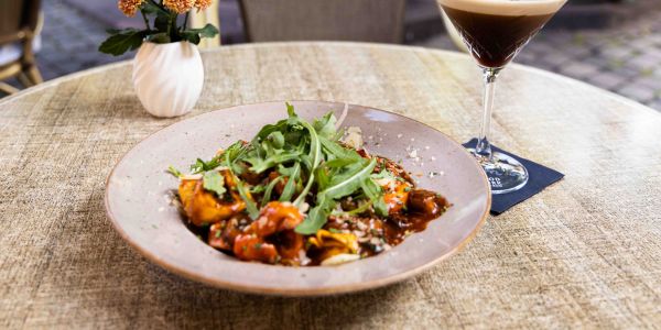 Ein runder Tisch im Freien mit einem tiefen Teller Pasta in dunkler Sauce, garniert mit Rucola, daneben eine Blumenvase und ein Espresso Martini im Cocktailglas.