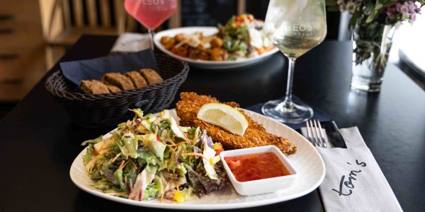 Ein paniertes Schnitzel mit Zitrone und Chilisauce neben einem großen Salat. Im Hintergrund stehen ein Brotkorb und Hauptspeisen. Daneben zwei bunte Cocktails.