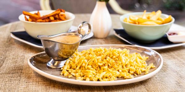 Ein runder Tisch im Freien mit verschiedenen Beilagen: ein Teller Spätzle mit einer Sauciere sowie Schalen mit Pommes frites und Süßkartoffelpommes.