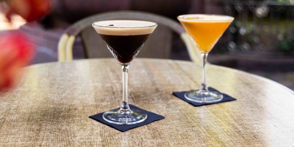 Ein runder Tisch im Außenbereich mit zwei Cocktails in Martinigläsern, jeweils auf einer dunklen Serviette.