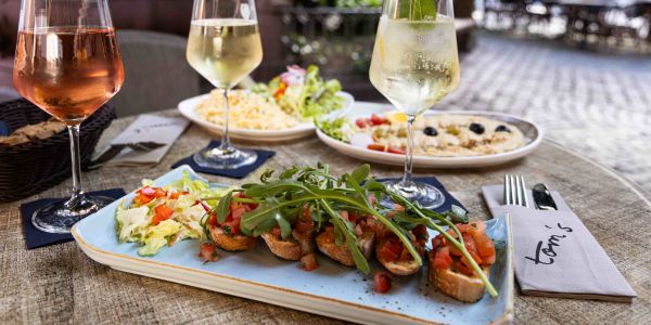 Ein gedeckter Tisch im Freien mit Gläsern für Rosé, Weißwein und einem Cocktail sowie Tellern mit Bruschetta, Salat und Hummus.
