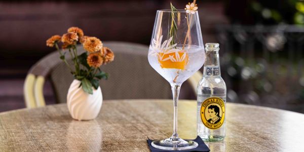 Ein runder Tisch im Außenbereich mit einer kleinen Blumenvase und einer Flasche Thomas Henry Tonic Water neben einem Gin Tonic im großen Weinglas, garniert mit einer Orangenschale auf einer dunklen Serviette.