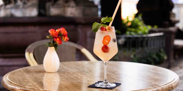 Ein runder Tisch im Außenbereich mit einer kleinen Blumenvase und einem Beeren-Cocktail mit Strohhalm und Minze auf einer dunklen Serviette.