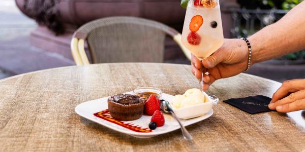 Ein runder Tisch im Außenbereich mit einem Dessertteller aus Lavakuchen, Eis und Erdbeeren, während eine Hand ein Cocktailglas mit Beeren dazustellt.