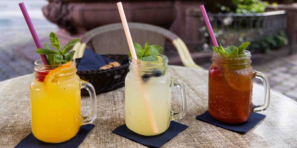 Ein runder Tisch im Außenbereich mit drei verschiedenen Limonaden in Henkelgläsern, garniert mit frischer Minze und Früchten, serviert auf dunklen Servietten.