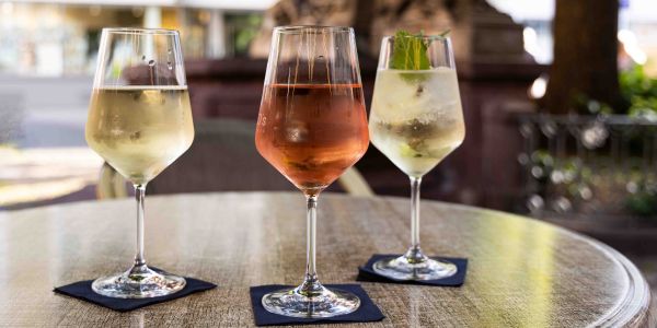 Ein runder Tisch im Außenbereich mit drei Weingläsern, gefüllt mit Weißwein, Rosé und einem Cocktail, jeweils serviert auf einer dunklen Serviette.