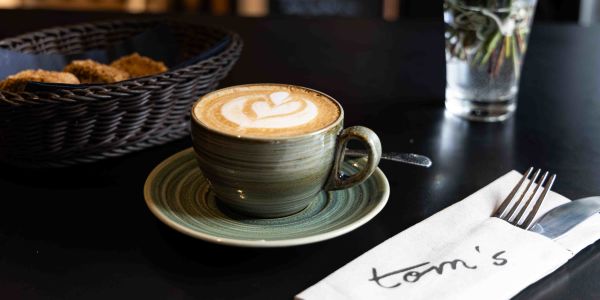 Eine Tasse Cappuccino mit kunstvollem Latte Art Herz auf einer passenden Untertasse. Daneben liegen ein Brotkorb und Besteck in einer "tom's"-Serviette.