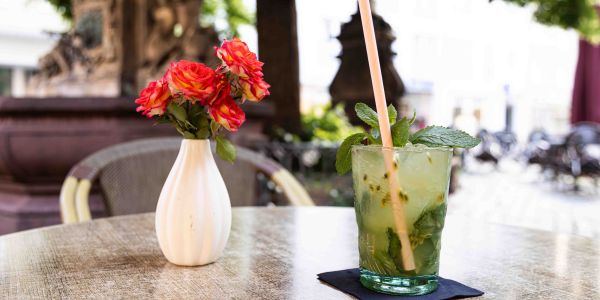 Ein runder Tisch im Außenbereich mit einer kleinen Vase voller roter Rosen und einem Glas Mojito mit Strohhalm und frischer Minze auf einer dunklen Serviette.