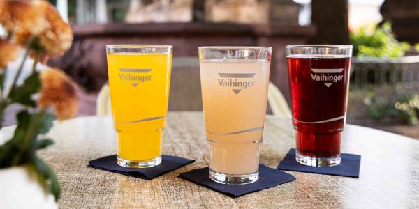 Ein runder Tisch im Freien mit drei Gläsern der Marke Vaihinger, gefüllt mit gelber, hellrosa und dunkelroter Saftschorle auf dunklen Servietten.